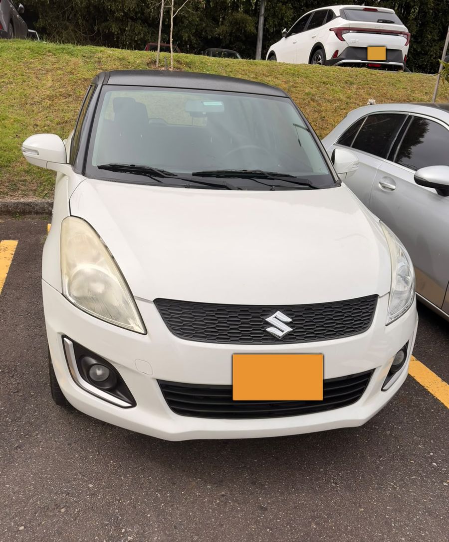 Suzuki Swift blanco sucio y con farolas amarillas, en un parqueadero